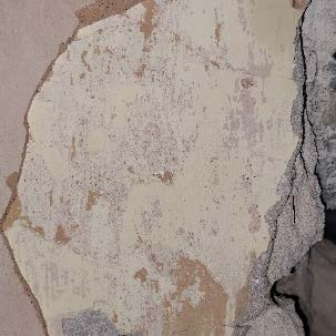 A close-up photograph showing a textured wall surface with peeling paint or plaster and a rough stone edge.