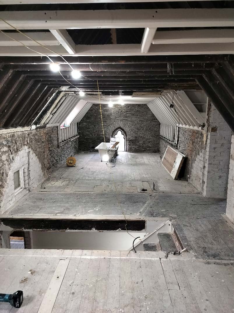 Interior photograph of a building under renovation featuring exposed stone walls, dark timber roof beams, and a bathtub placed in the center of the room.