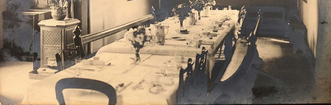A sepia-toned photograph showing the interior of a room featuring a long dining table set with flowers and chairs, alongside a stove.