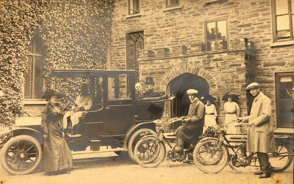 A vintage sepia-toned photograph showing a group of people posing with an early automobile and motorcycles in front of a historic stone building covered in ivy.