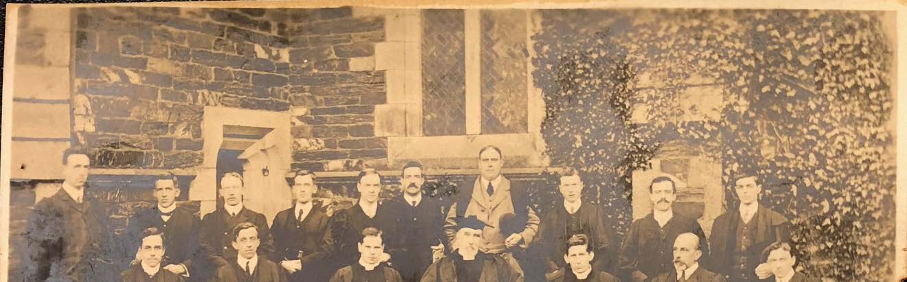 A vintage sepia-toned photograph showing a large group of men posing in front of a stone building with ivy-covered walls and boarded windows.