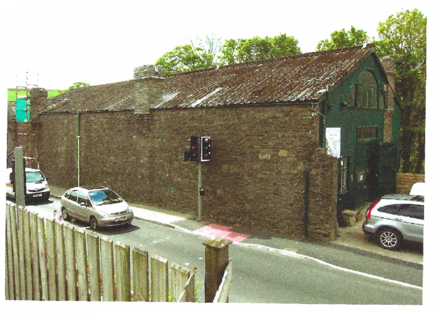 A photograph showing a long, single-story stone building with a weathered corrugated roof situated alongside a road with parked cars and a traffic light.