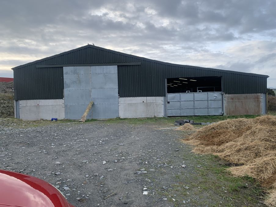 A large agricultural building with dark corrugated metal cladding and concrete lower walls, featuring large sliding doors and situated in a gravel yard with hay bales.