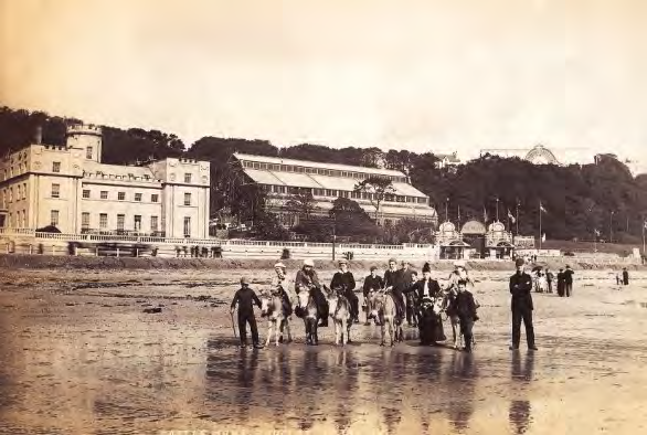 A vintage sepia-toned photograph depicting a beach scene with people riding ponies, set against a backdrop of large coastal buildings and a hillside.