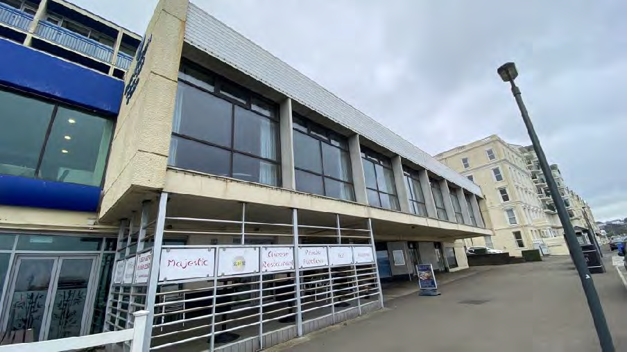 A street-level photograph of a multi-story commercial building with a concrete facade and large windows, featuring signage for a restaurant and bar.