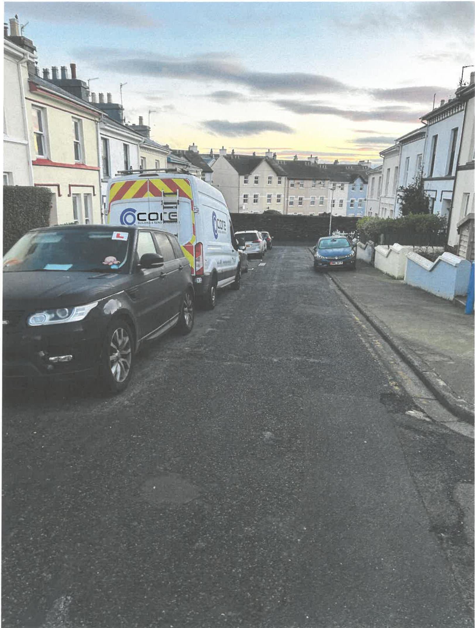 A street-level photograph showing a residential road lined with terraced houses and parked vehicles, including a black SUV and a white van.