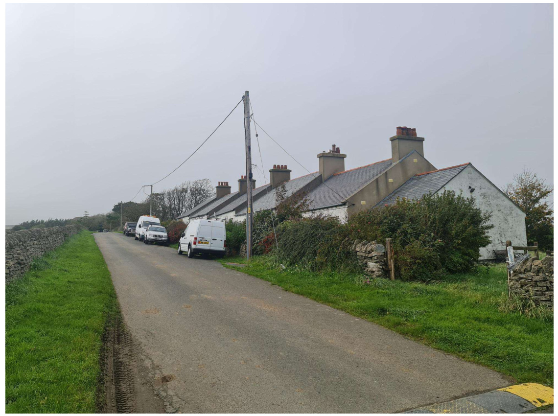 A photograph showing a rural road lined with a stone wall on the left and a row of white-walled farm buildings on the right, with several white vans parked along the side.