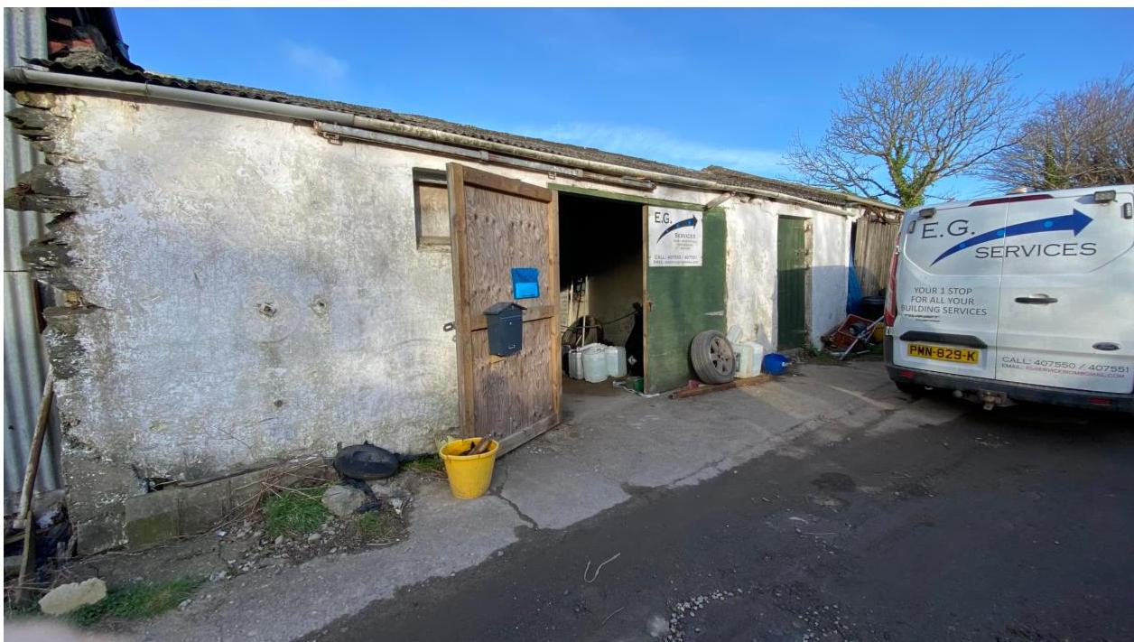 A photograph showing the exterior of a long, white-walled outbuilding or workshop with large wooden doors and a parked service van.