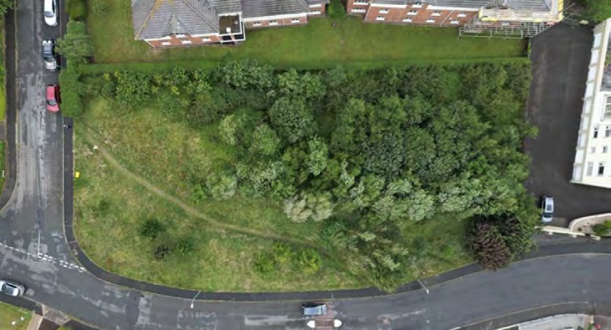 An aerial photograph showing a green, wooded plot of land surrounded by roads and residential buildings, serving as a site location view.