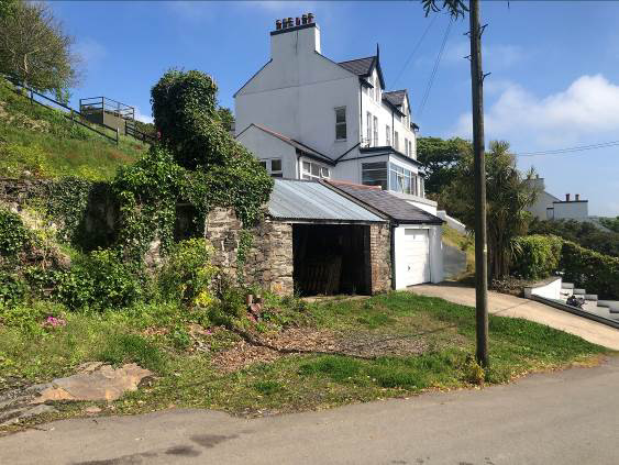 A photograph showing a derelict stone garage structure with a corrugated roof in the foreground, situated below a white three-story house on a sloping site.