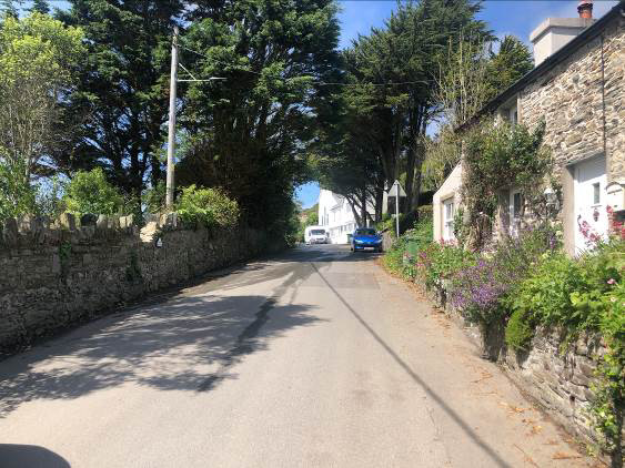 A street-level photograph showing a paved road lined with stone walls and traditional stone cottages under a clear blue sky.