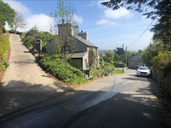 A street-level photograph showing a residential property on a sloping site with a driveway, stone walls, and surrounding vegetation.