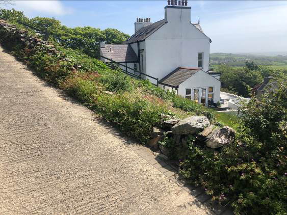 A photograph showing a white detached house situated on a slope with a stone wall and paved road in the foreground, overlooking a rural landscape.