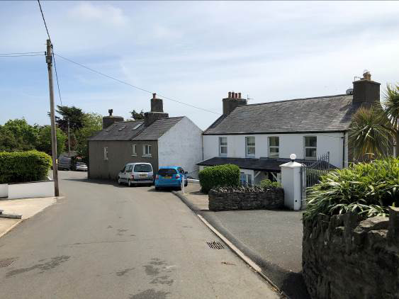A street-level photograph showing a residential scene with a white house and a smaller grey outbuilding or garage. The image captures the existing site context including parked cars, stone walls, and surrounding green...
