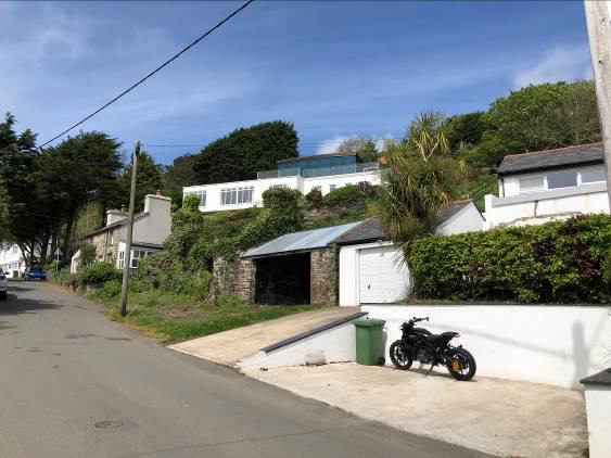 A street-level photograph showing a driveway with a motorcycle and bin, leading to a pair of garages (one open, one closed) on a sloping residential site.
