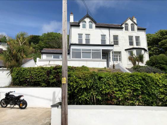 A street-level photograph of a large white detached house situated on a hillside, with a motorcycle parked in the foreground and a wooden utility pole.