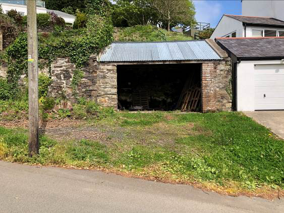 A photograph showing a derelict stone garage with a corrugated roof situated on a sloping site next to a modern white garage.
