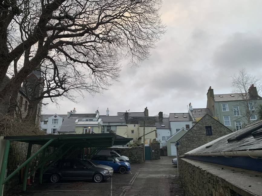 A photograph showing a residential street scene with a green carport and parked cars in the foreground, and a row of traditional terraced houses in the background.