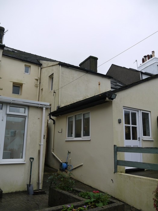 The image displays the rear exterior of a cream-colored terraced house with white windows and a small patio area. A green wooden fence and garden planters are visible in the foreground.