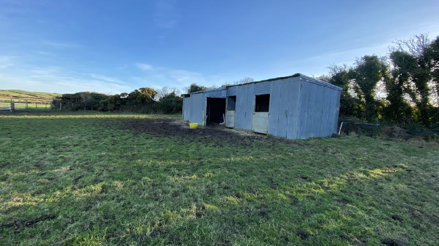 A photograph showing a corrugated metal stable building situated in a grassy field with trees and rolling hills in the background.