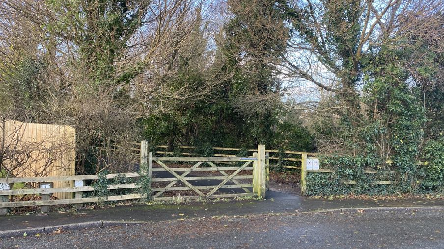 A photograph showing a wooden gate and fence at a property entrance, surrounded by dense trees and vegetation.
