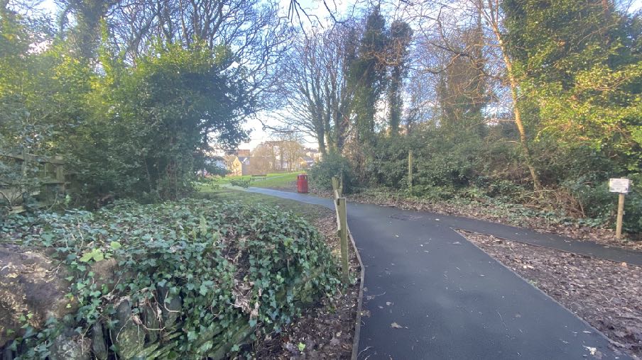 A photograph showing a paved path winding through a wooded area with a stone wall covered in ivy and houses visible in the background.