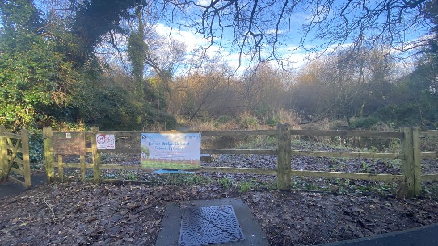 A photograph showing a wooden fence with a community notice attached, set against a background of trees and foliage with a metal grate in the foreground.