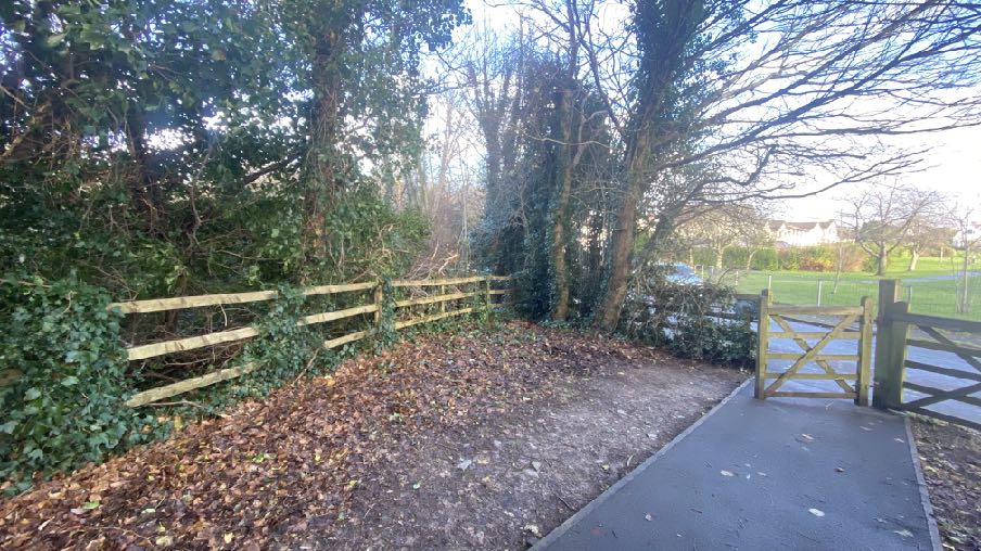 A photograph showing a wooden fence and gate at the edge of a property, with a paved path leading to a gate and trees in the background.