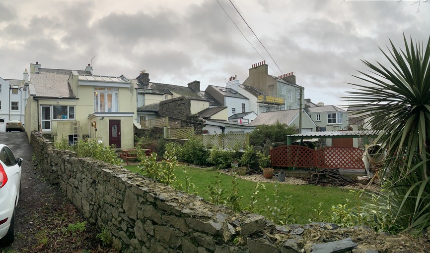 A street-level photograph showing a residential property with a stone boundary wall, garden, and neighboring houses under a cloudy sky.