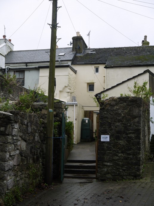 A street-level photograph showing the entrance to a white-walled house with a slate roof, accessed via a paved path through stone walls and a wooden gate.