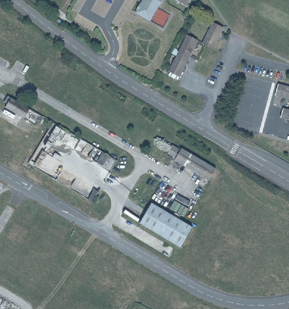 An aerial photograph showing an industrial or commercial site adjacent to a road, featuring buildings, paved yards, and parking areas.