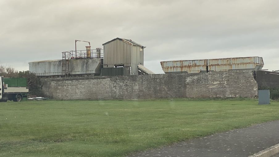 A photograph showing an industrial site with large metal hoppers and structures behind a brick wall, with a truck visible on the left.