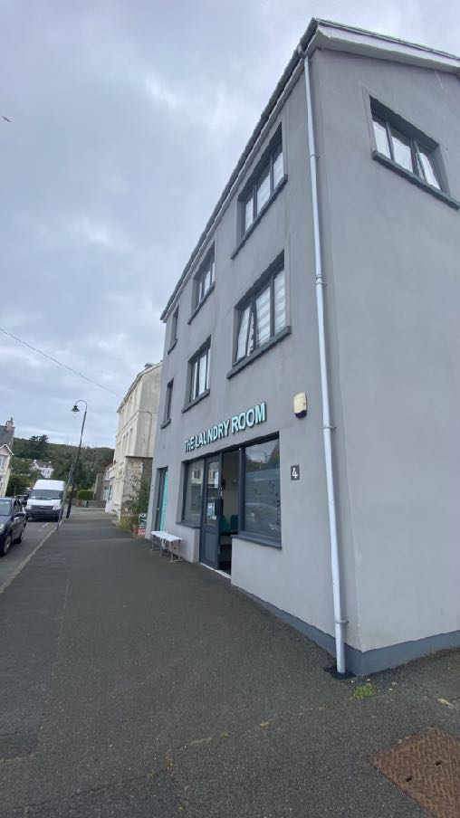 A street-level photograph showing the exterior facade of a grey, three-story commercial building with a sign reading 'THE LAUNDRY ROOM'.