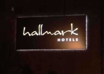 A night-time photograph of an illuminated rectangular sign reading 'hallmark HOTELS' mounted on a dark brick wall.