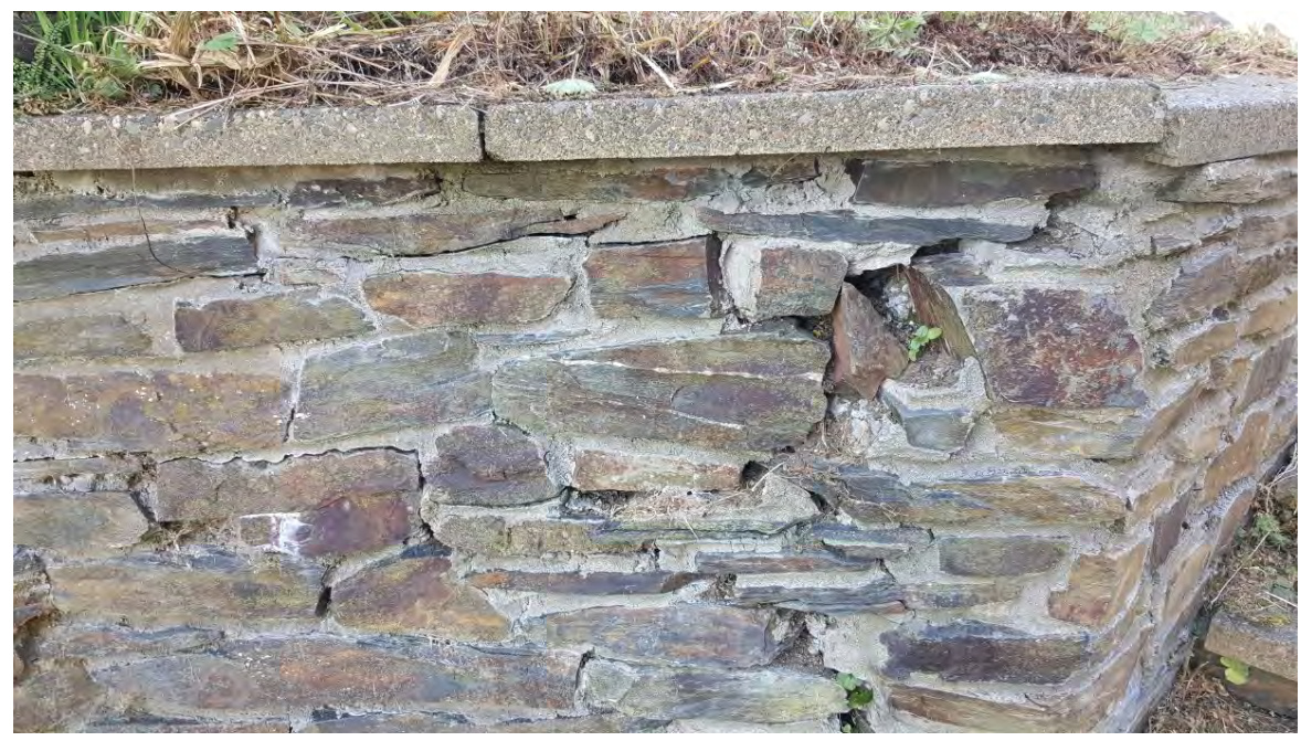 A close-up photograph of a weathered stone retaining wall with a concrete coping stone on top.