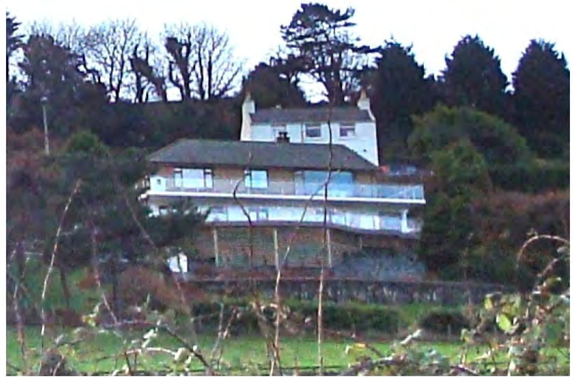 A photograph showing a large, modern detached house built into a sloping, wooded hillside with visible retaining walls.