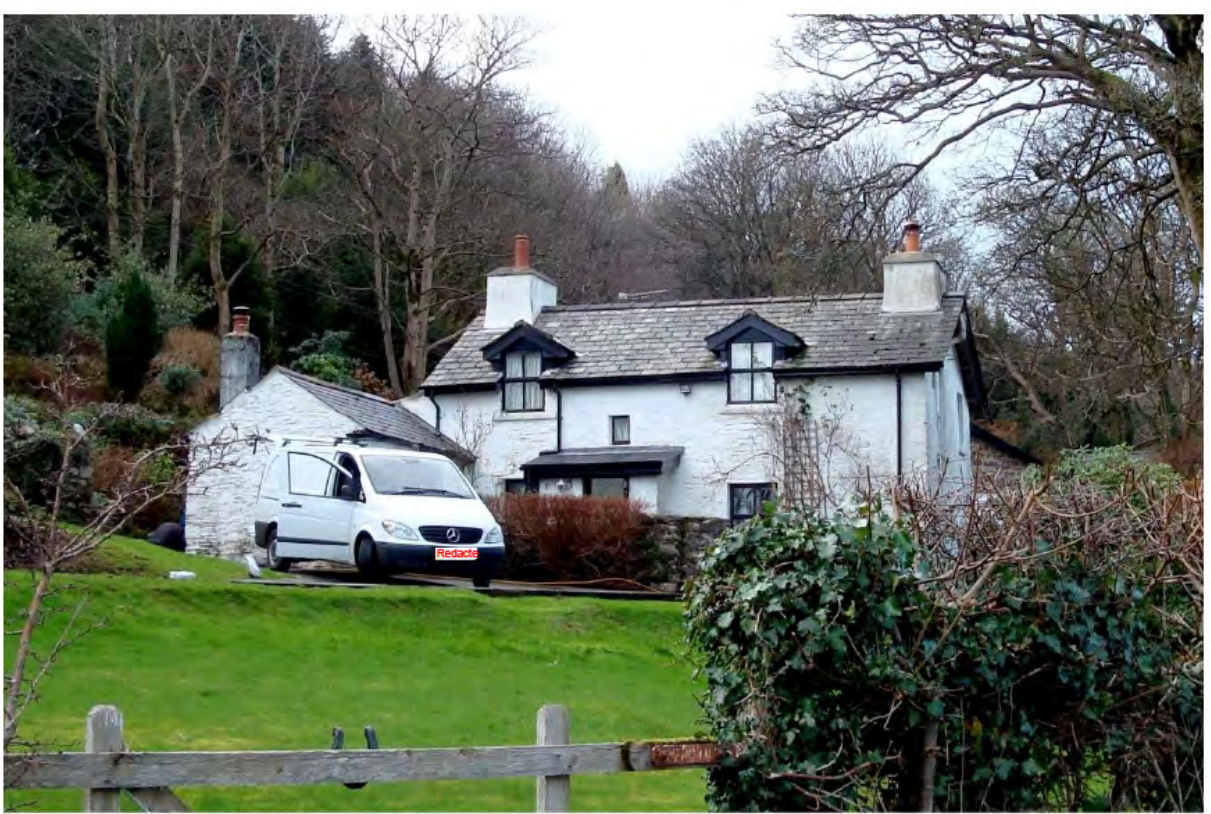 A photograph of a white, two-story detached house with a slate roof situated on a grassy slope in a rural setting.