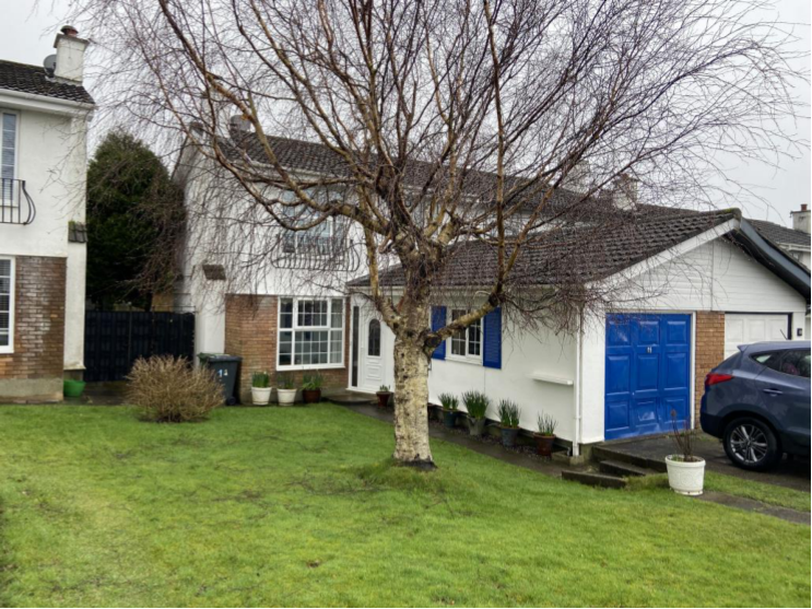 A photograph showing the front exterior of a detached residential property with a large tree in the garden and a garage with blue doors.