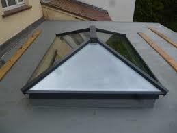 A close-up photograph of a triangular glass roof lantern installed on a flat grey roof, with wooden planks nearby.