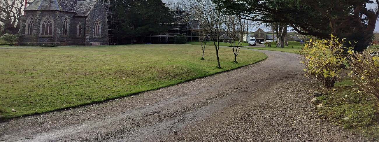 A photograph showing a gravel driveway curving through grassy grounds towards a stone building with Gothic windows and scaffolding.