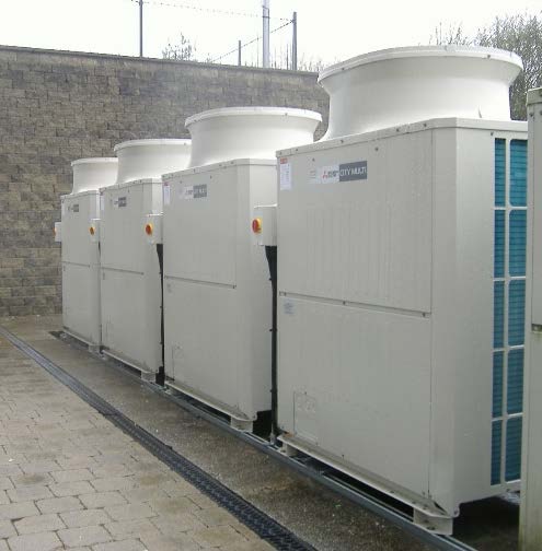 A row of large white external plant units, likely HVAC equipment, installed on a paved surface next to a stone wall.
