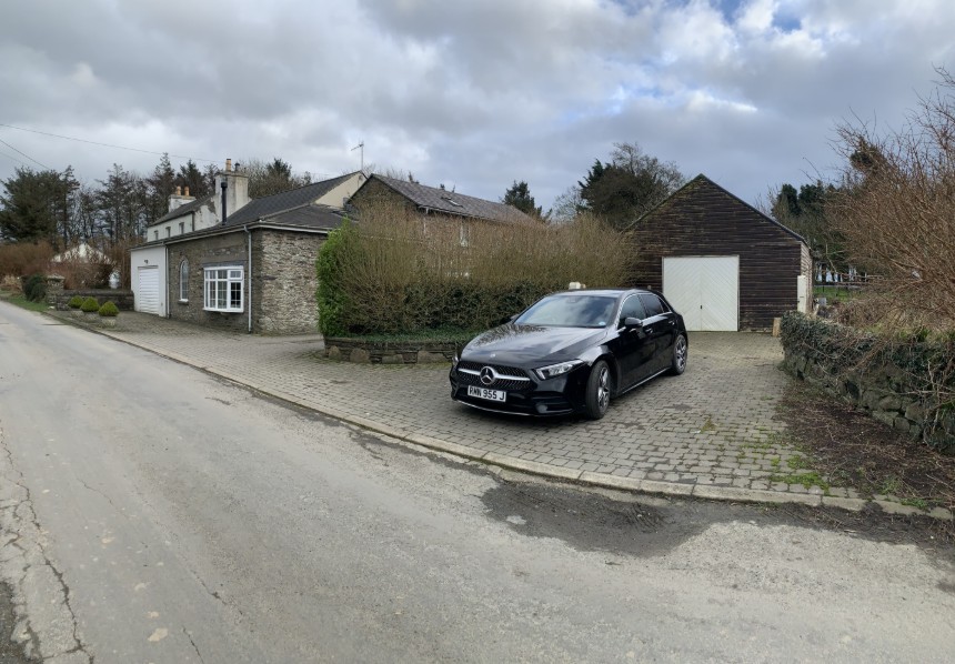 A photograph showing a stone house with an attached garage and a separate wooden garage/workshop to the right, with a black car parked on the paved driveway.