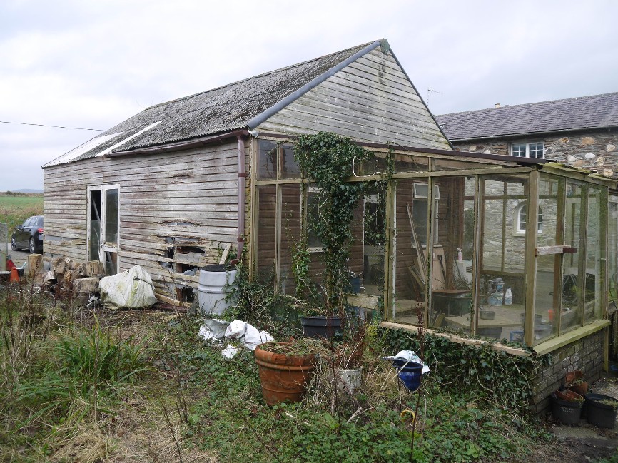 A weathered wooden outbuilding or garage with an attached glass lean-to structure, showing signs of disrepair and overgrown surroundings.