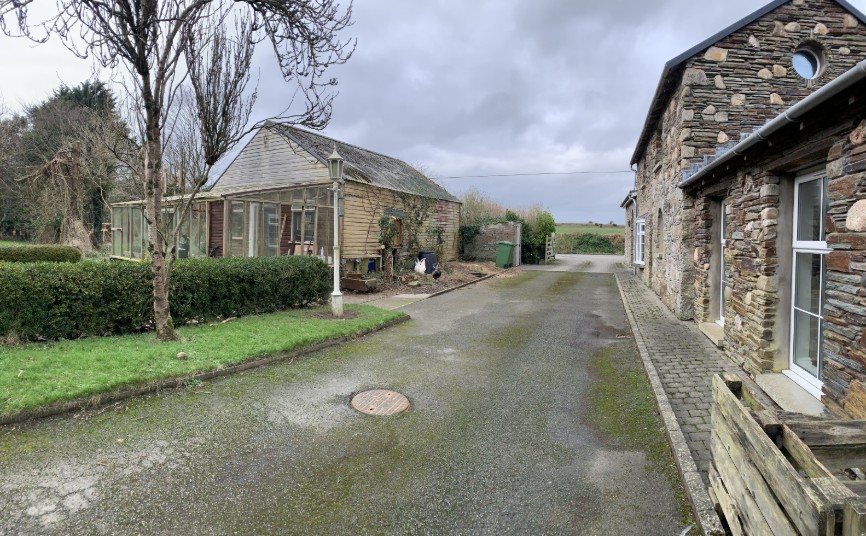 A photograph showing a rural driveway with a stone house on the right and an existing wooden garage/workshop with a glass porch on the left.