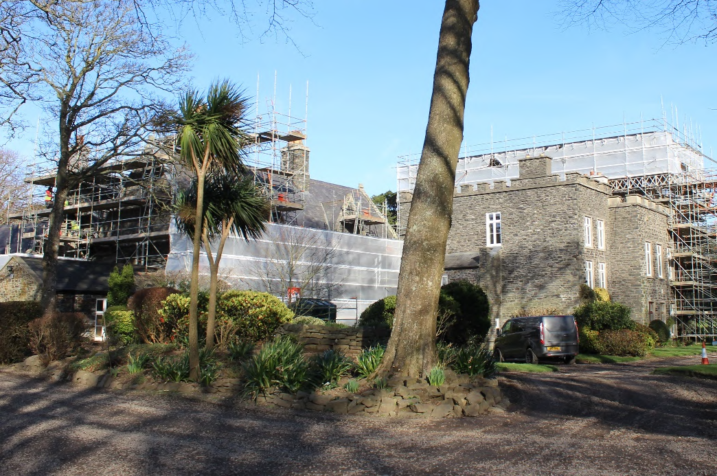 A photograph showing a large stone building covered in scaffolding and protective sheeting, viewed from a gravel driveway with landscaping in the foreground.