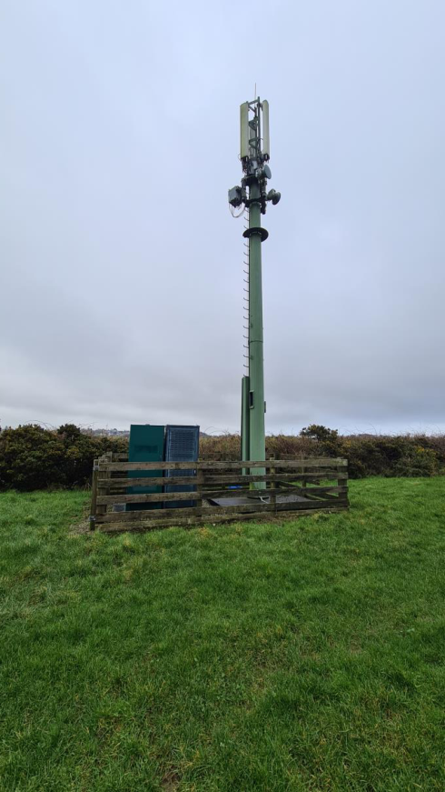 A photograph showing an existing green telecommunications monopole mast with ground equipment cabinets enclosed by a wooden fence in a grassy field.