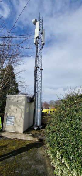A photograph showing a tall telecommunications lattice mast with antennas and a beige ground equipment cabinet situated in a green, semi-rural setting.