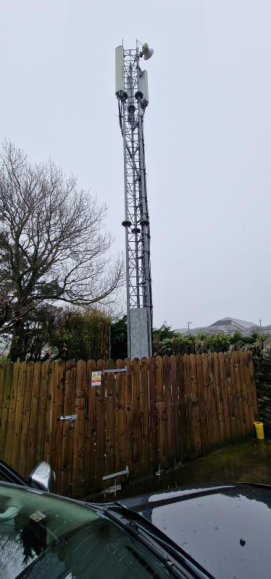 A photograph showing a tall telecommunications lattice mast with antennas, situated behind a wooden fence in a rural setting.