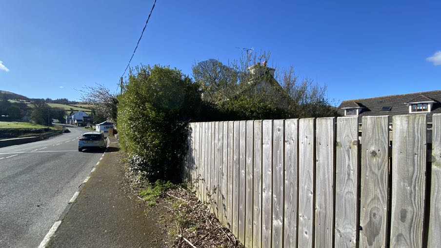 A street-level photograph showing a wooden fence running along a roadside with a car and houses visible in the background.