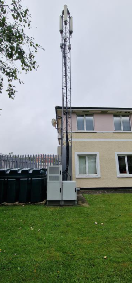 A photograph showing a tall lattice telecommunications tower with panel antennas and ground equipment cabinets situated next to a two-story building.